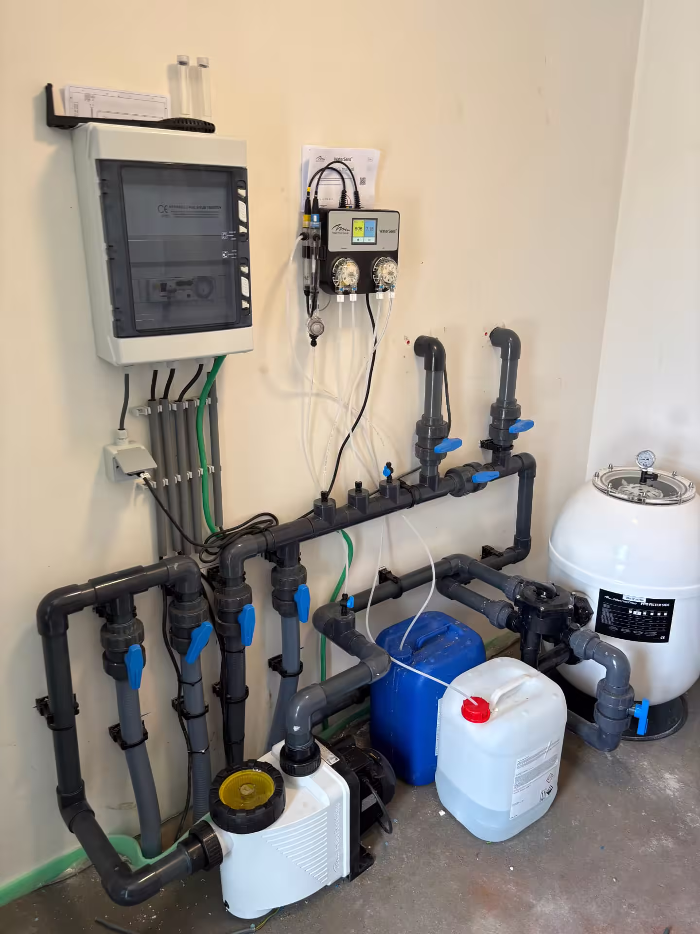 Technische ruimte met PVC-leidingwerk, zandfilter, zwembadpomp en WaterSens-doseersysteem tegen een lichte muur